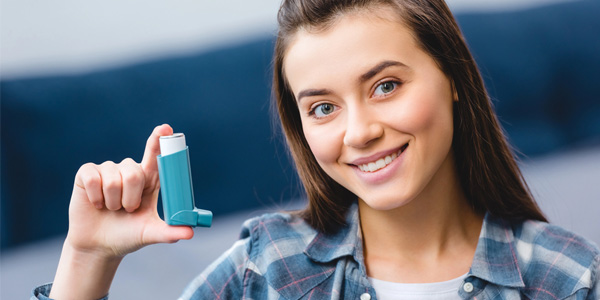 Person hält einen blauen Asthma-Inhalator in der rechten Hand vor dem Körper. Darstellung kann auf Beratung zur korrekten Anwendung und Inhalationstechnik hinweisen.